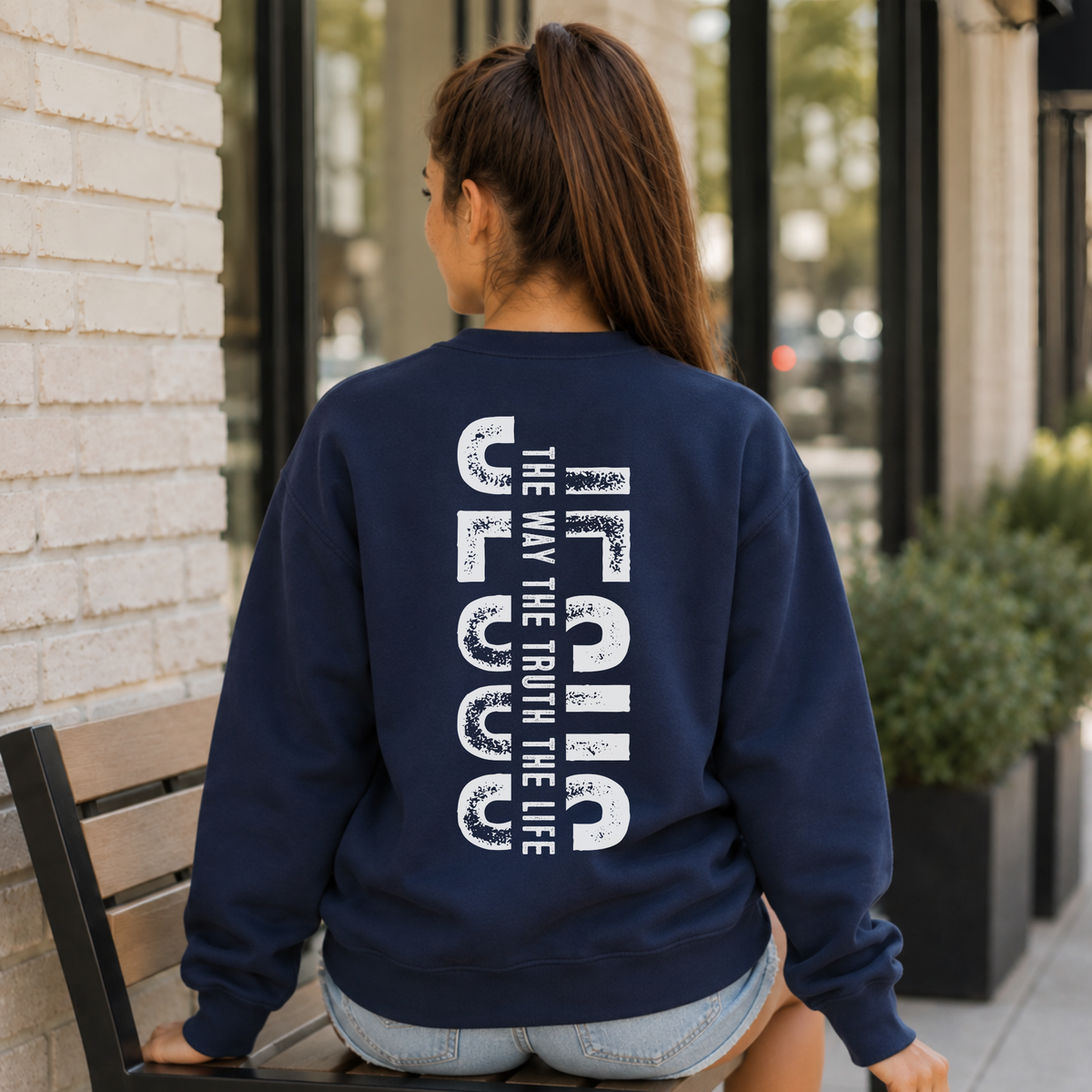 Woman shown from behind wearing a navy crewneck sweatshirt with vertical white JESUS lettering and “The Way, The Truth, The Life” text.