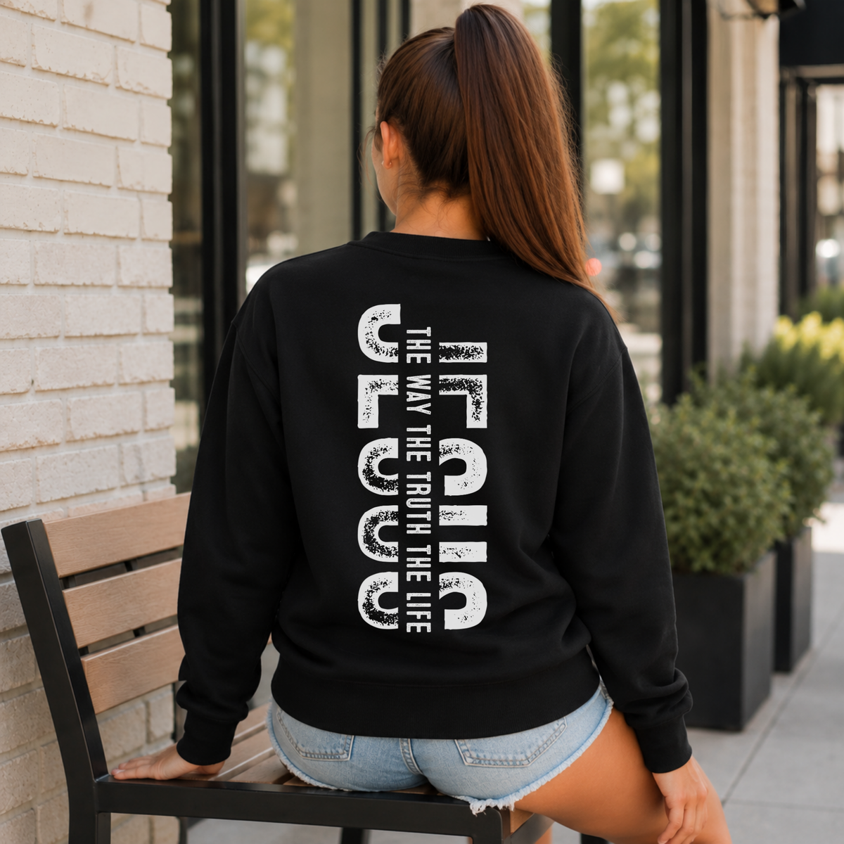 Woman shown from behind wearing a black crewneck sweatshirt with vertical white JESUS lettering and “The Way, The Truth, The Life” text.