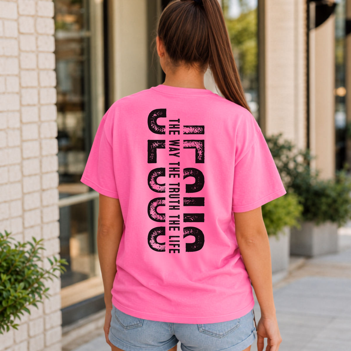 Woman shown from behind wearing a bright pink T-shirt with vertical black JESUS lettering and “The Way, The Truth, The Life” text.