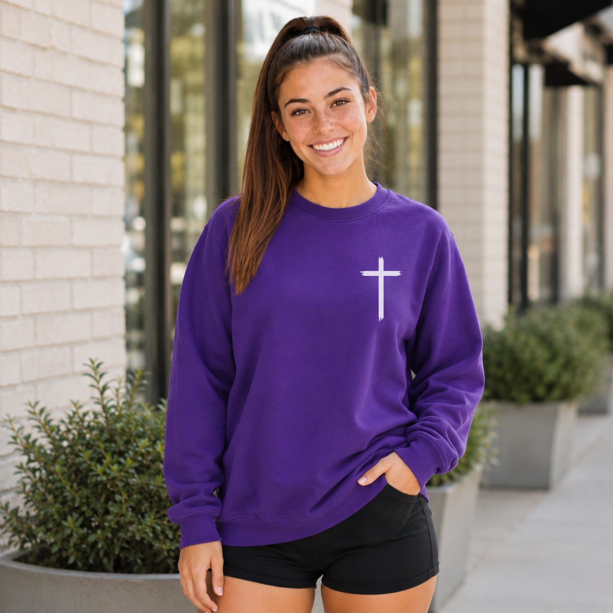 Woman wearing a purple crewneck sweatshirt with a white cross on the front chest outside a storefront.