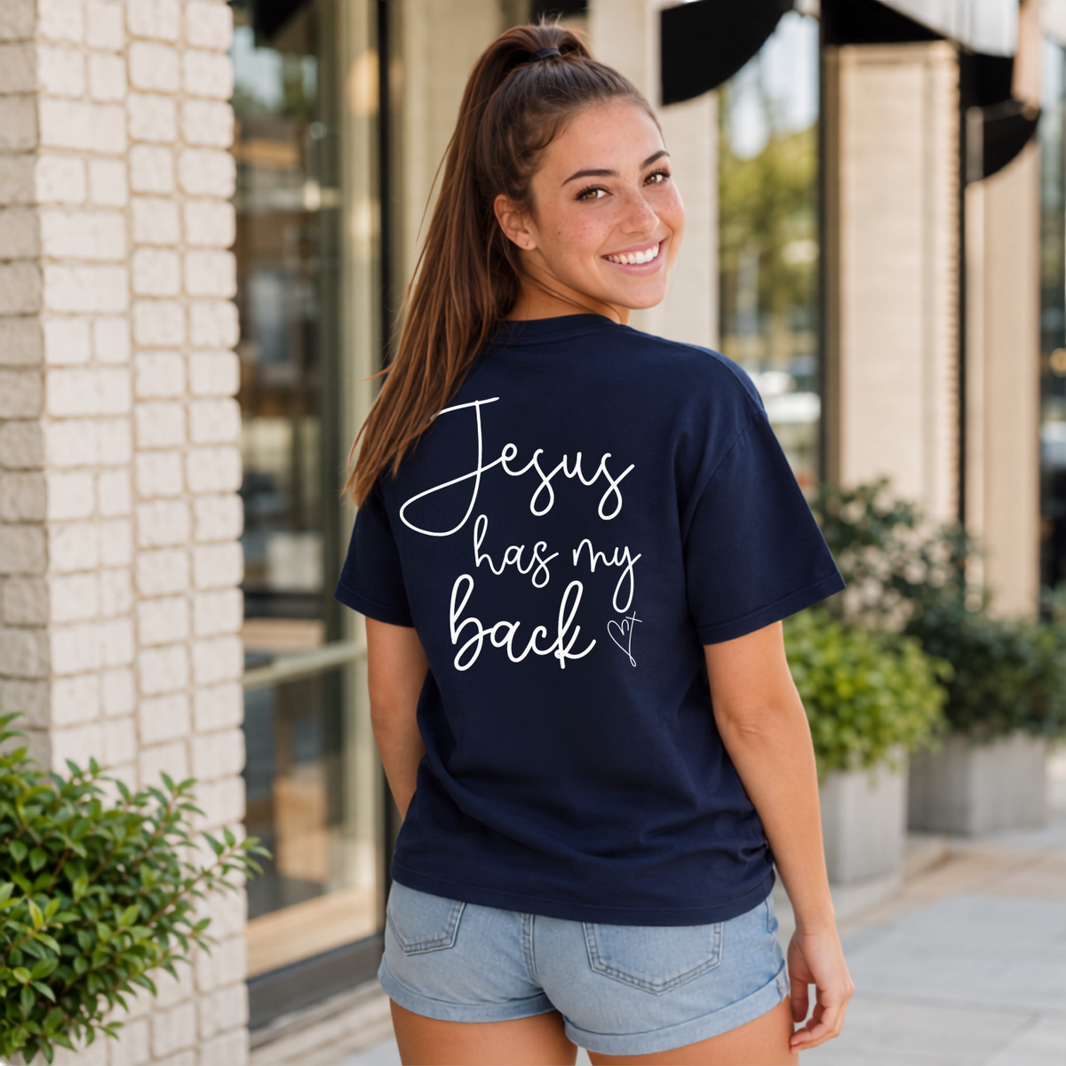 Back view of a woman wearing a navy Jesus Has My Back T-shirt with white script lettering and a heart cross graphic.