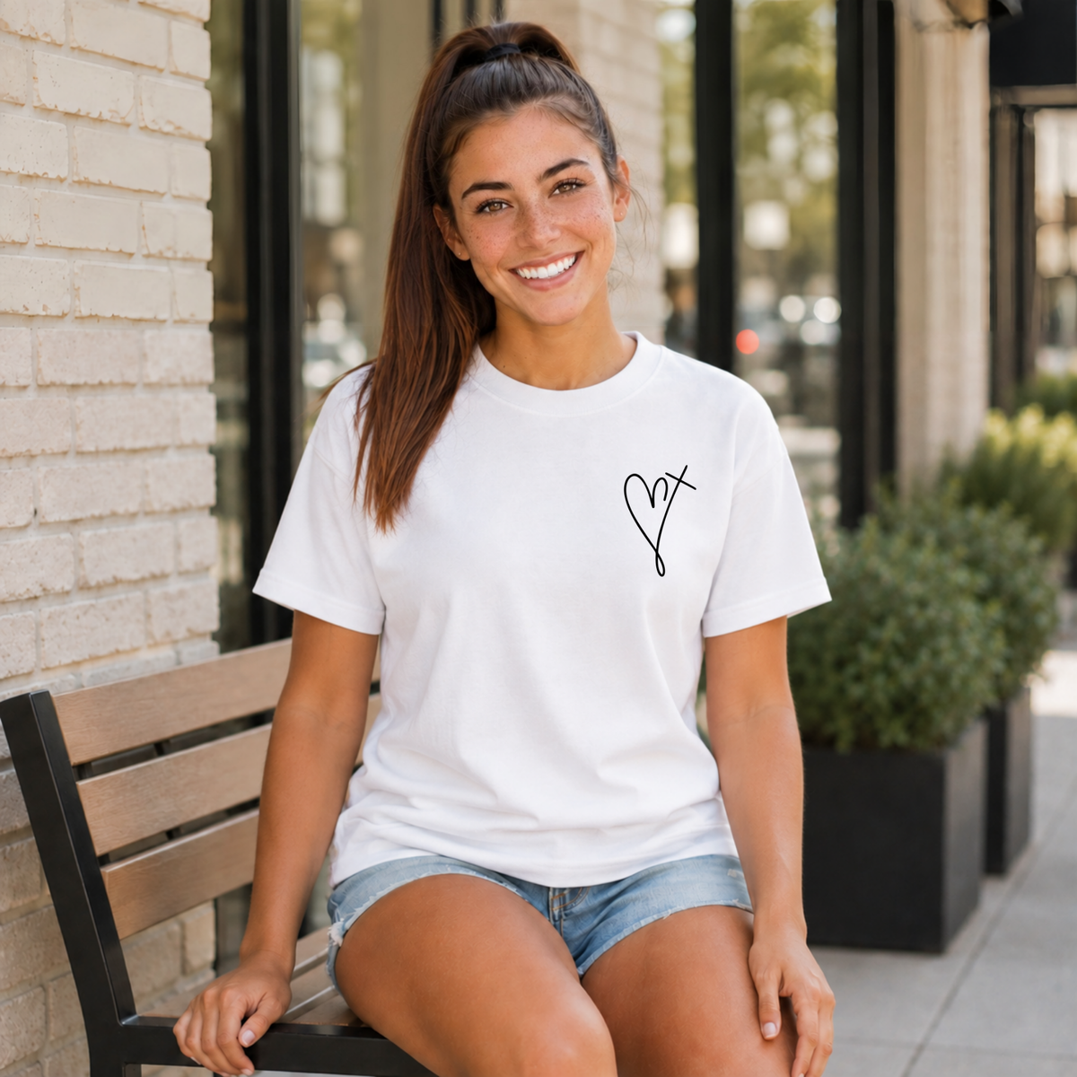 Woman wearing a white Jesus Has My Back T-shirt with a small heart cross graphic on the front chest while seated outside.