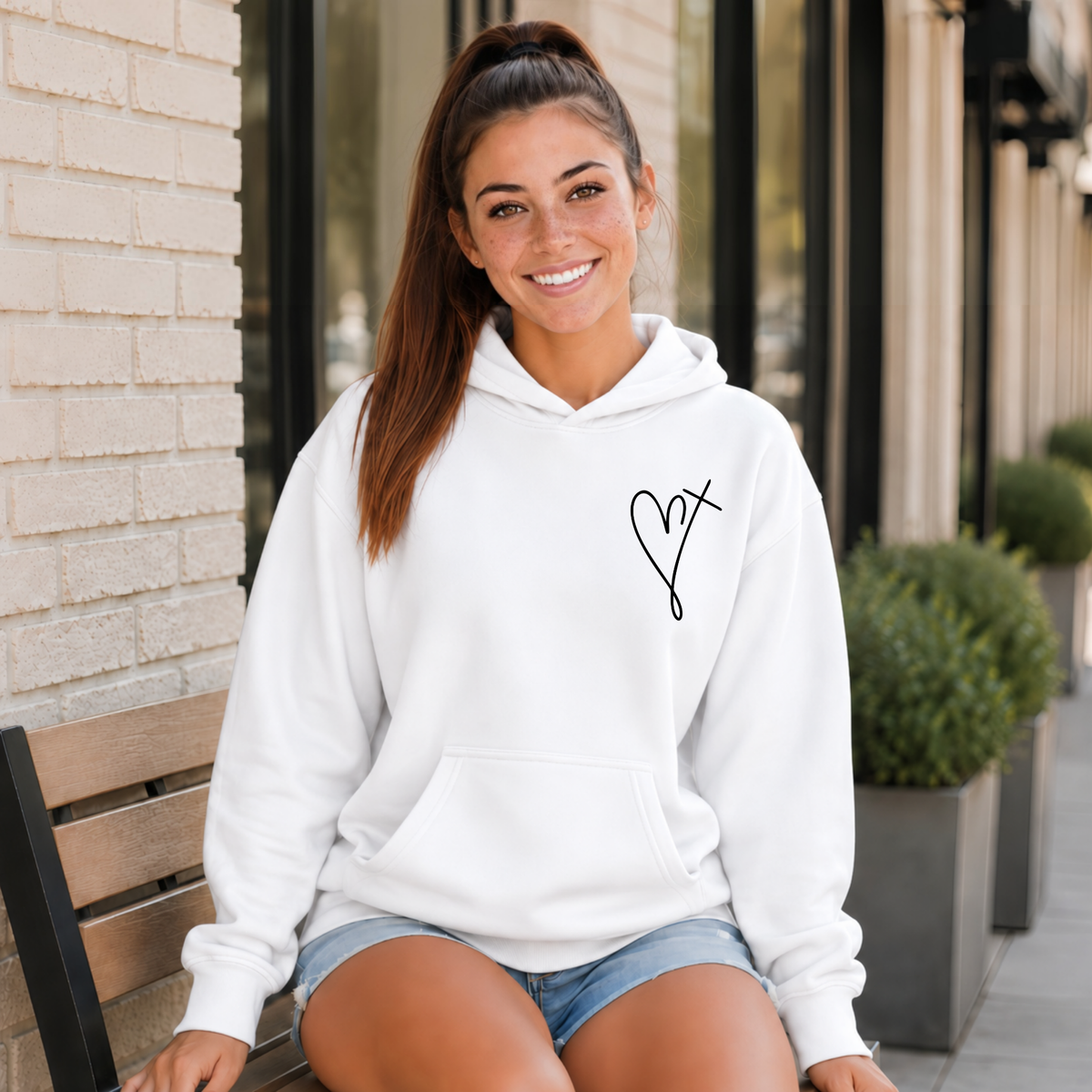 Woman wearing a white Jesus Has My Back hoodie with a small heart cross graphic on the front chest outside a storefront.