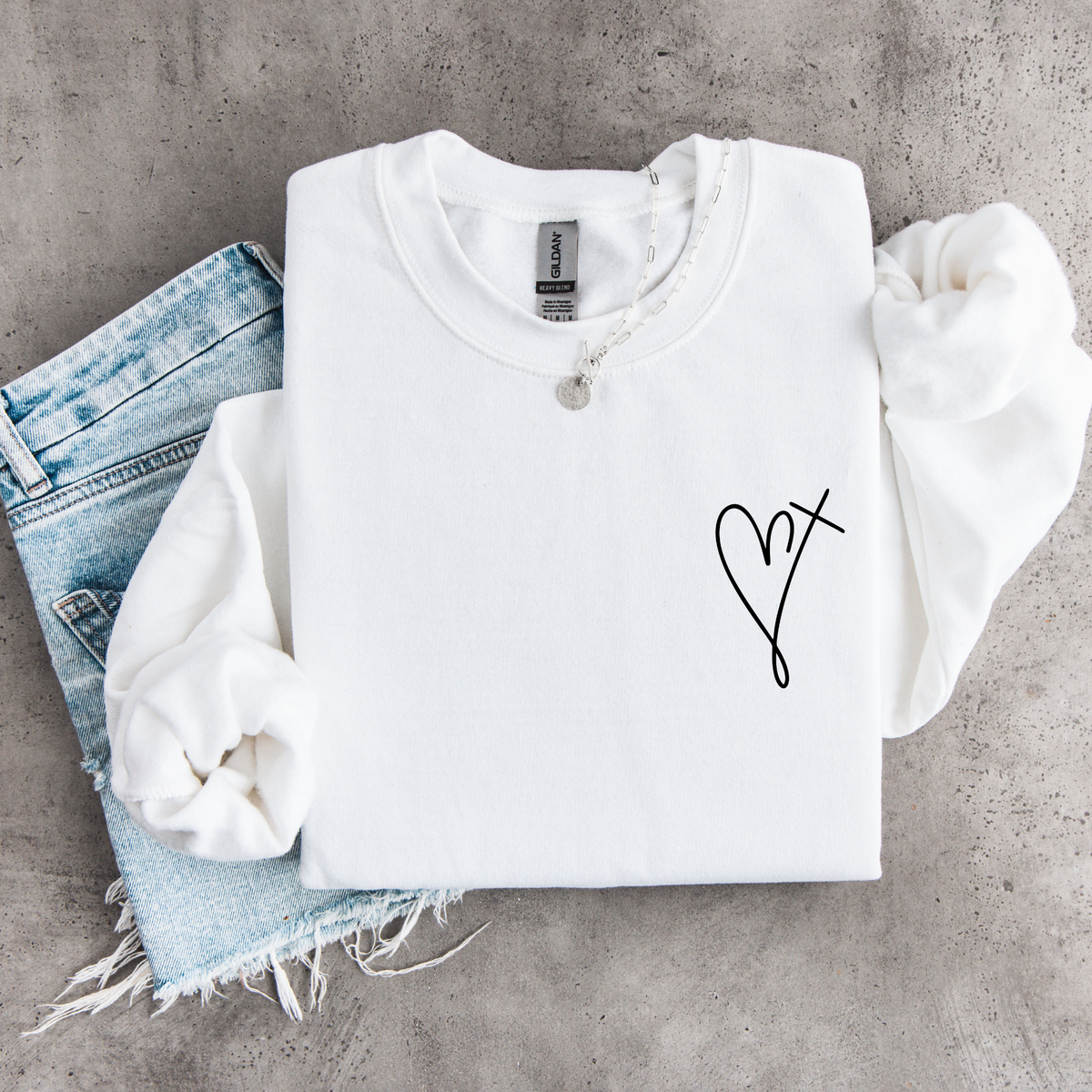 White youth sweatshirt flat lay with a small heart cross graphic on the front chest, styled with denim.
