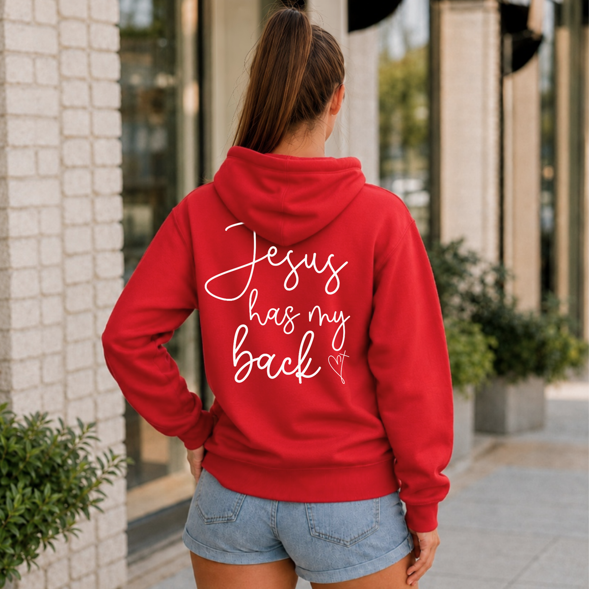 Back view of a woman wearing a red Jesus Has My Back hoodie with white script lettering and a heart cross graphic.
