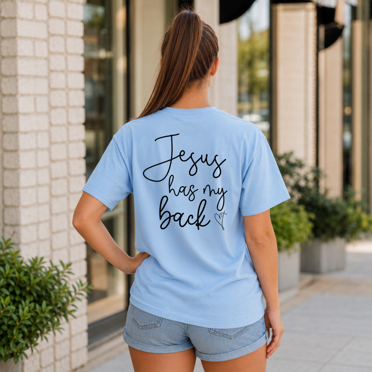 Back view of a woman wearing a light blue Jesus Has My Back T-shirt with black script lettering and a heart cross graphic.