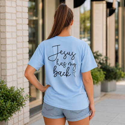 Back view of a woman wearing a light blue Jesus Has My Back T-shirt with black script lettering and a heart cross graphic.