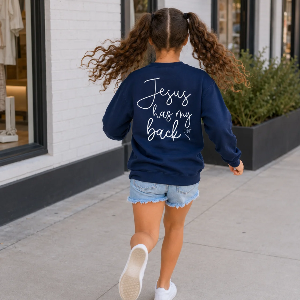 Back view of a girl wearing a navy youth sweatshirt with Jesus Has My Back script lettering and a heart cross graphic outside a storefront.