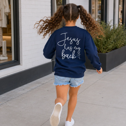 Back view of a girl wearing a navy youth sweatshirt with Jesus Has My Back script lettering and a heart cross graphic outside a storefront.