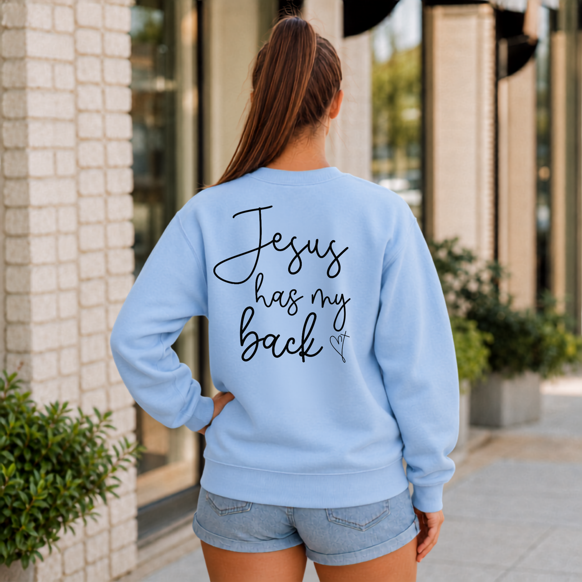 Back view of a woman wearing a light blue Jesus Has My Back crewneck sweatshirt with black script lettering and a heart cross graphic.
