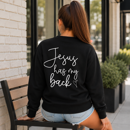 Back view of a woman wearing a black Jesus Has My Back crewneck sweatshirt with white script lettering and a heart cross graphic.