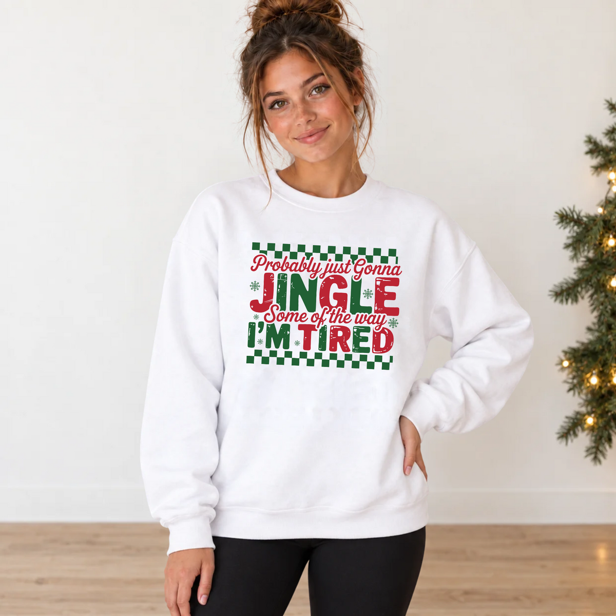 Woman in a white sweatshirt with red and green Jingle Some of the Way I'm Tired Christmas graphic beside a lit evergreen garland.