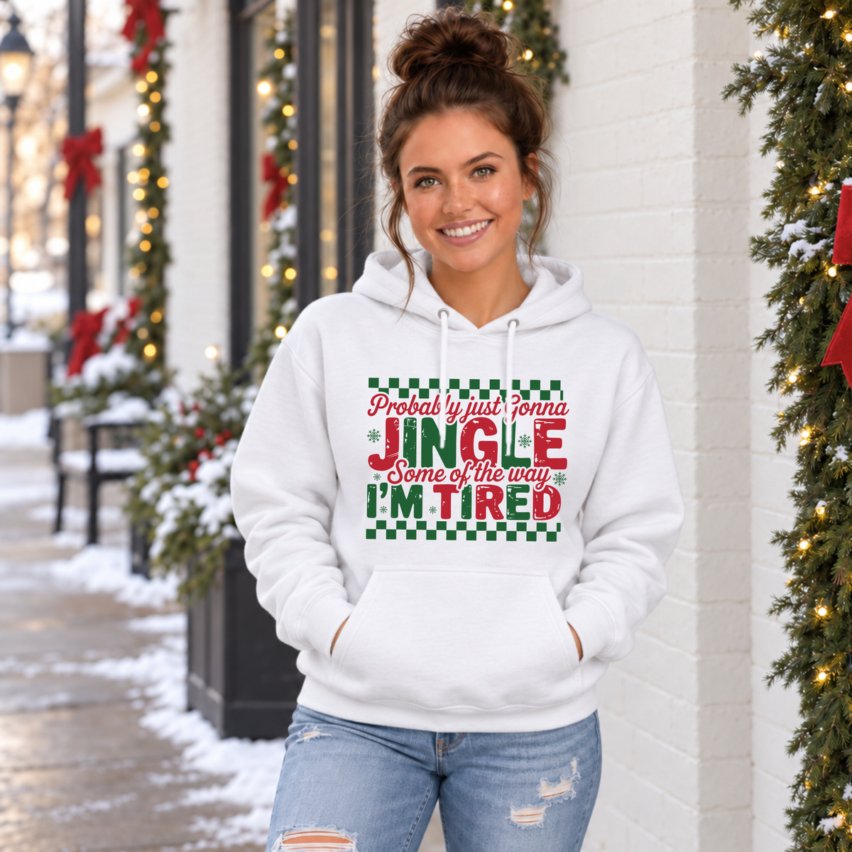 Woman wearing a white hoodie with red and green Probably Just Gonna Jingle Some of the Way I'm Tired Christmas design outside a snowy storefront