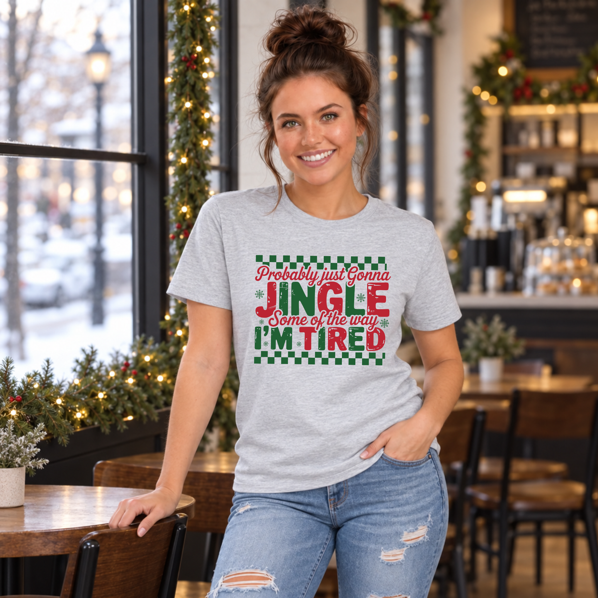 Woman wearing a sport gray T-shirt with red and green Probably Just Gonna Jingle Some of the Way I'm Tired Christmas design in a café