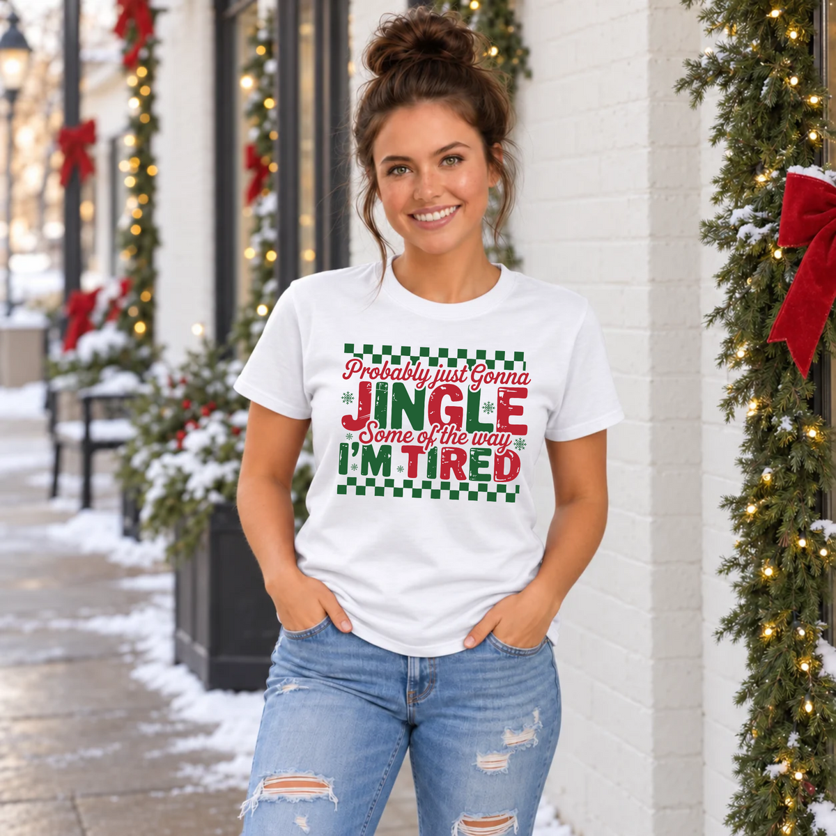 Woman wearing a white T-shirt with red and green Probably Just Gonna Jingle Some of the Way I'm Tired Christmas design outside a snowy, decorated storefront.