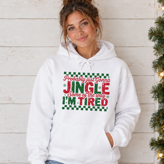 Woman in a white hoodie with tied drawstrings and red and green Jingle Some of the Way I'm Tired Christmas graphic beside a lit evergreen garland.