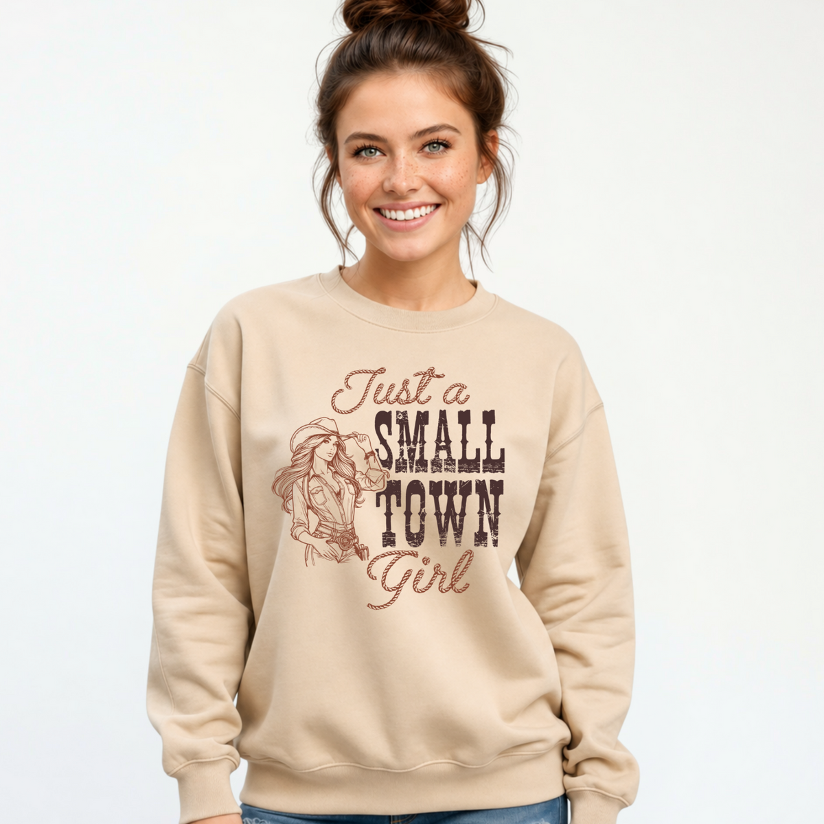 Woman wearing a sand crewneck sweatshirt with the “Just a Small Town Girl” western cowgirl graphic, photographed against a clean light studio background.