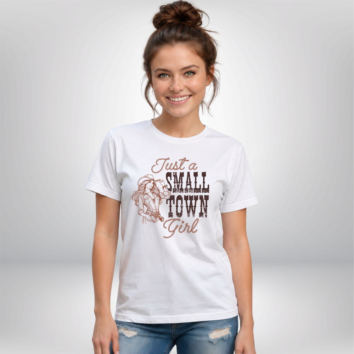 Woman wearing a white T-shirt with the “Just a Small Town Girl” cowgirl graphic, shown in a simple studio-style setting with a light background.