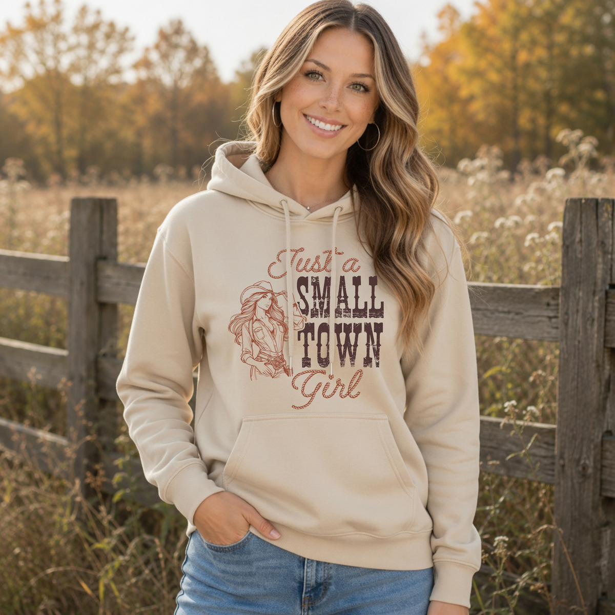 Woman wearing a sand hoodie with the “Just a Small Town Girl” vintage cowgirl graphic outdoors beside a rustic wooden fence in an autumn field.
