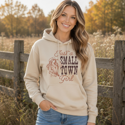 Woman wearing a sand hoodie with the “Just a Small Town Girl” vintage cowgirl graphic outdoors beside a rustic wooden fence in an autumn field.