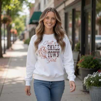 Woman walking downtown in a white crewneck sweatshirt featuring the “Just a Small Town Girl” western cowgirl design, with storefronts and flowers in the background.