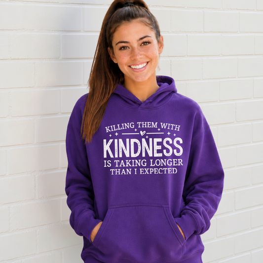 Woman wearing a purple hoodie with white Killing Them With Kindness lettering against a white brick wall.