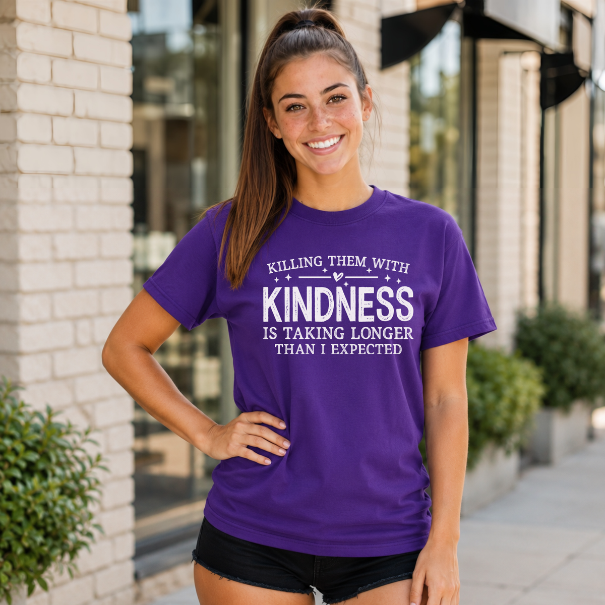Woman wearing a purple T-shirt with white Killing Them With Kindness lettering outside a storefront.
