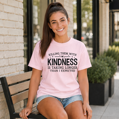 Woman seated outside wearing a light pink T-shirt with black Killing Them With Kindness lettering.
