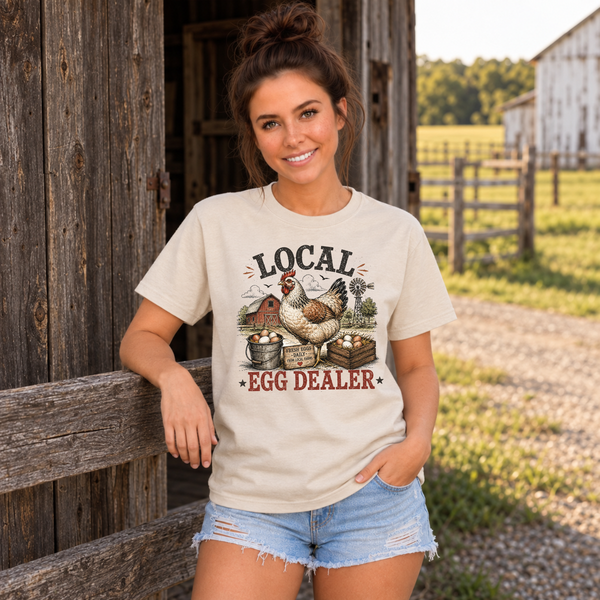 Woman wearing a sand Local Egg Dealer T-shirt leaning on a wooden farm fence.