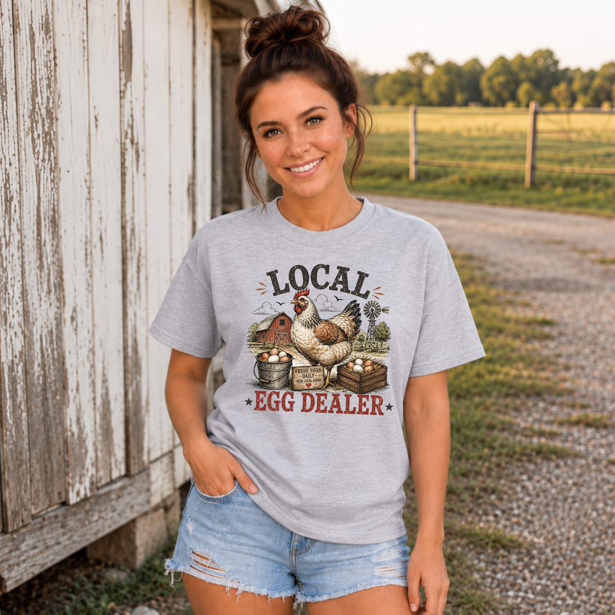 Woman wearing a sports gray Local Egg Dealer T-shirt beside a weathered white barn.