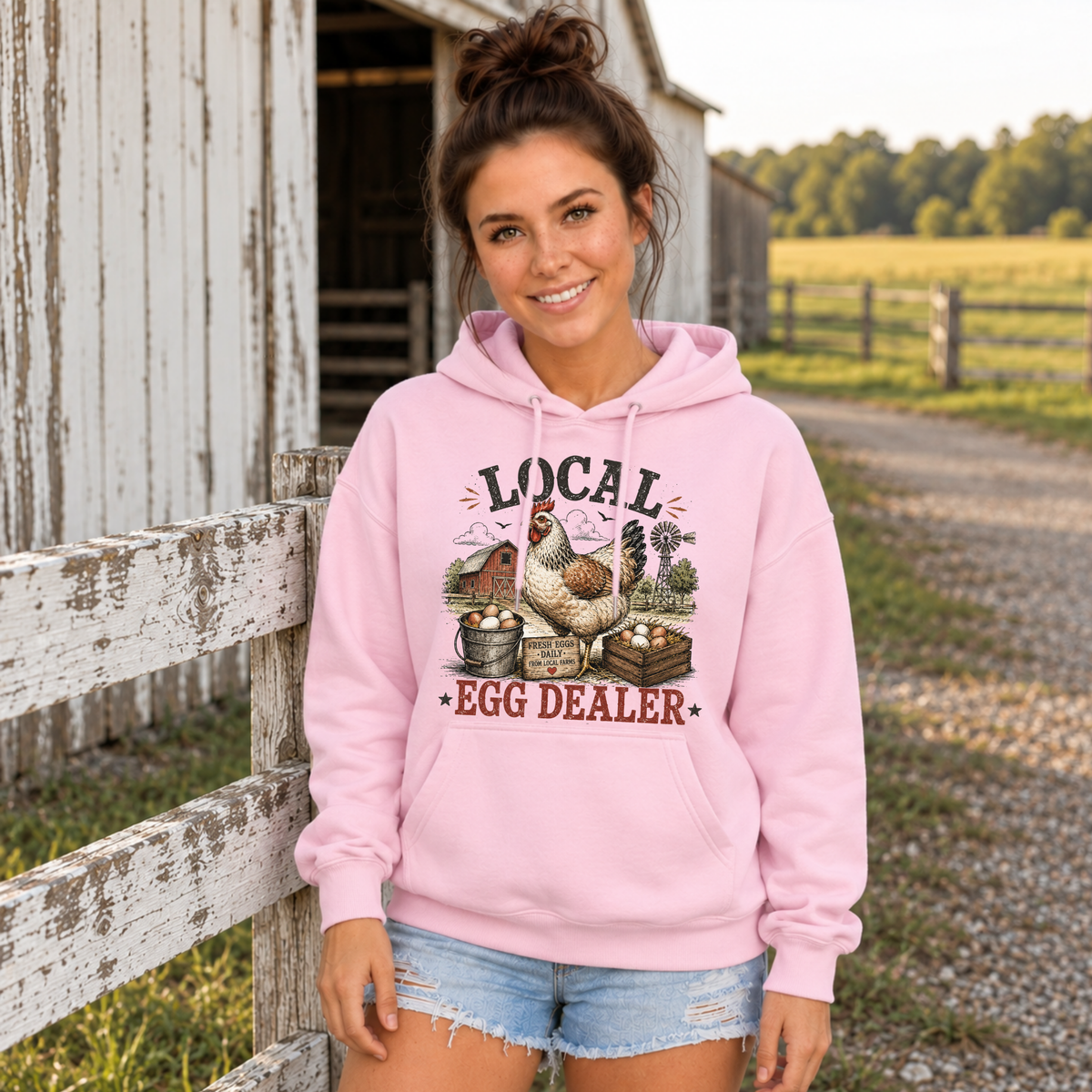 Woman in a light pink Local Egg Dealer hoodie leaning by a white farm fence.