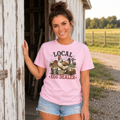 Woman wearing a light pink Local Egg Dealer T-shirt beside a weathered barn doorway.
