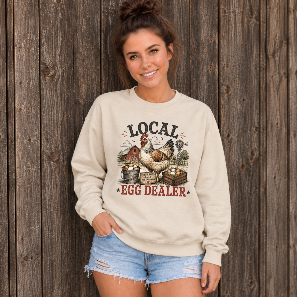Woman in a sand Local Egg Dealer sweatshirt against a dark wooden barn wall.