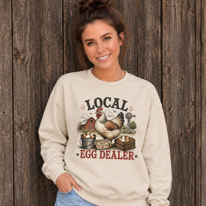 Woman wearing a sand Local Egg Dealer sweatshirt with hen, eggs, barn, and windmill graphic against a weathered wood wall.