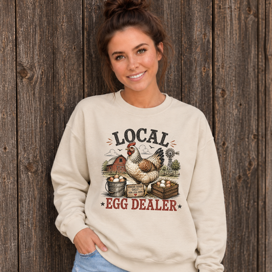 Woman wearing a sand Local Egg Dealer sweatshirt with hen, eggs, barn, and windmill graphic against a weathered wood wall.
