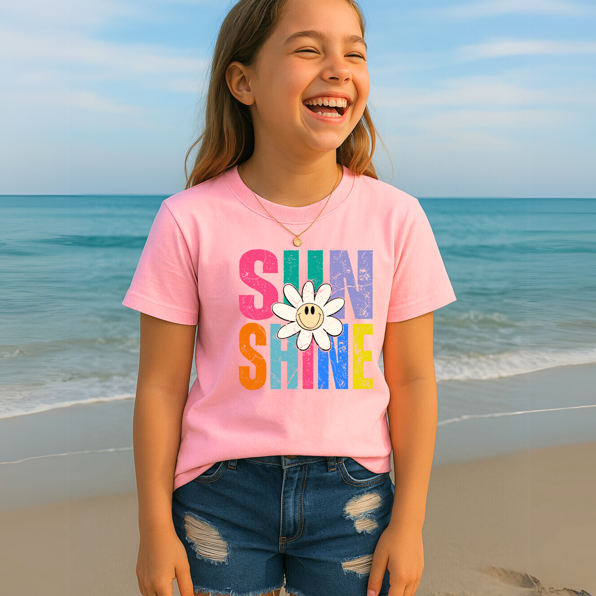 Laughing girl on the beach in a pastel pink Sunshine T-shirt featuring a cheerful daisy and vibrant rainbow design

