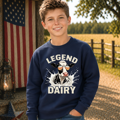 A smiling boy wears a navy crewneck sweatshirt with a “LEGEND DAIRY” graphic showing a black-and-white cow in sunglasses splashing through milk, in a rustic outdoor setting with an American flag.