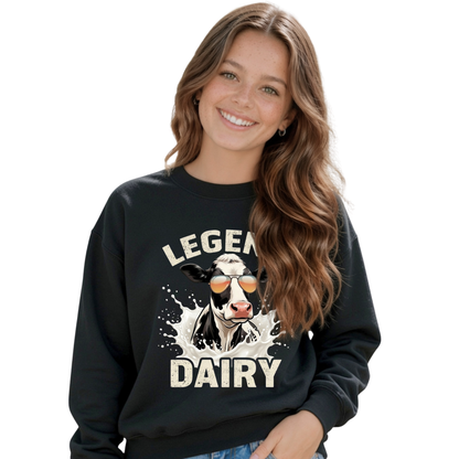 A smiling young girl wears a black crewneck sweatshirt with a “LEGEND DAIRY” graphic showing a black-and-white cow in sunglasses splashing through milk, against a white background.
