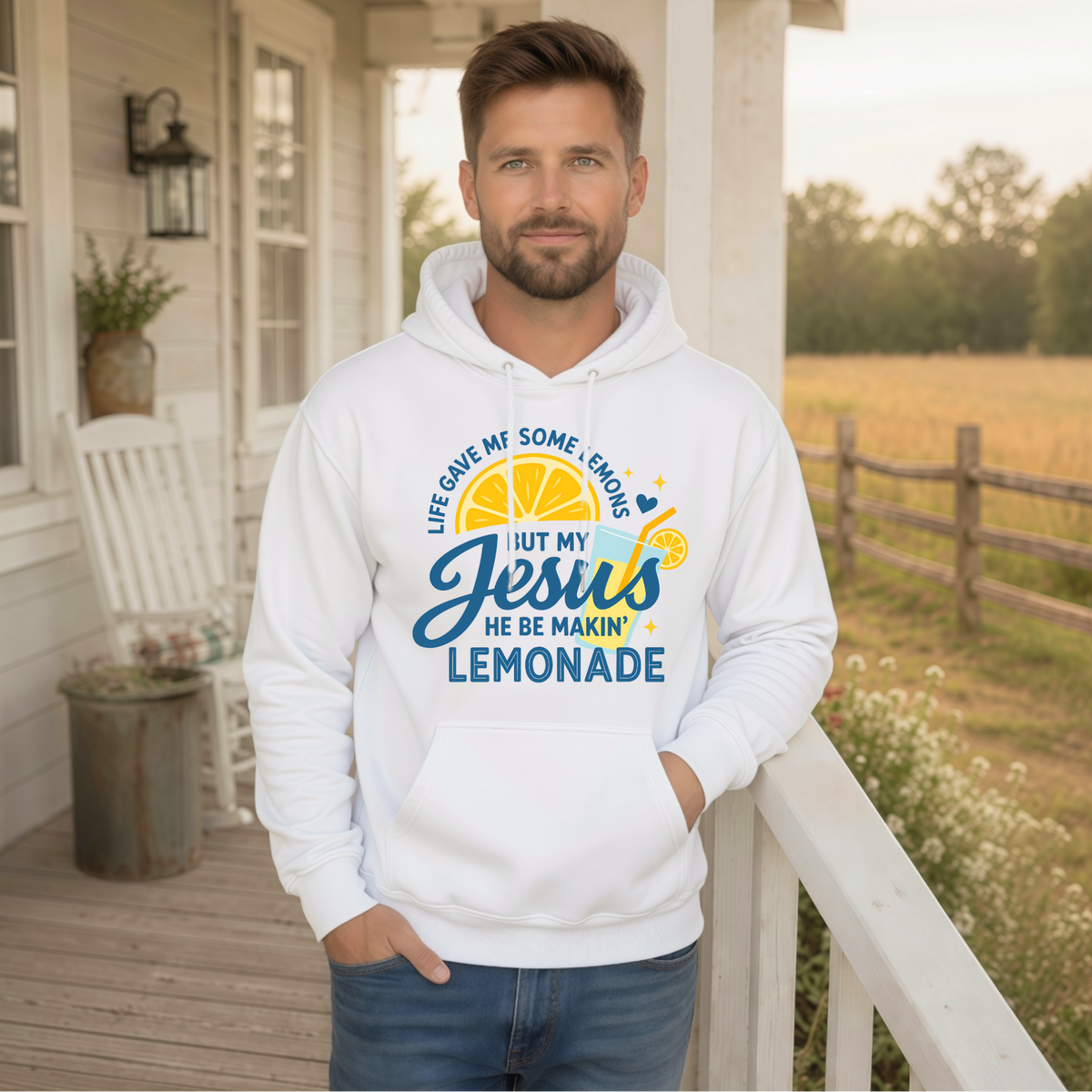 Man wearing a white hoodie printed with the Jesus Be Makin’ Lemonade lemon and lemonade cup graphic on a farmhouse porch. 