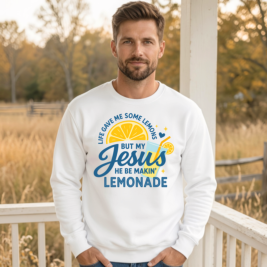 Man wearing a white crewneck sweatshirt with the Jesus Be Makin’ Lemonade graphic featuring blue lettering, lemons and a lemonade drink design outdoors.