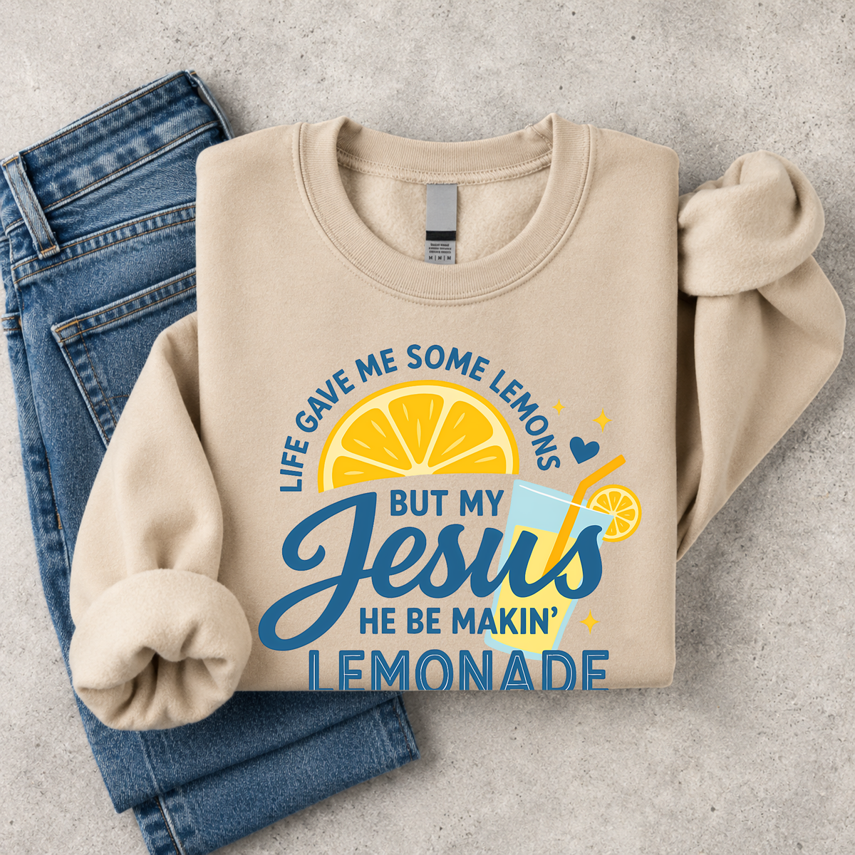 Folded sand crewneck sweatshirt flat lay with the Life Gave Me Some Lemons, But My Jesus He Be Makin’ Lemonade graphic beside blue jeans.