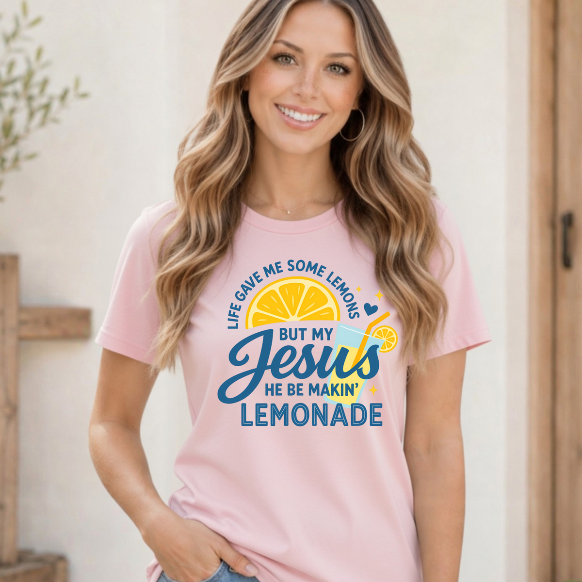 Woman wearing a light pink T-shirt with the Life Gave Me Some Lemons, But My Jesus He Be Makin’ Lemonade faith graphic on a bright neutral background.