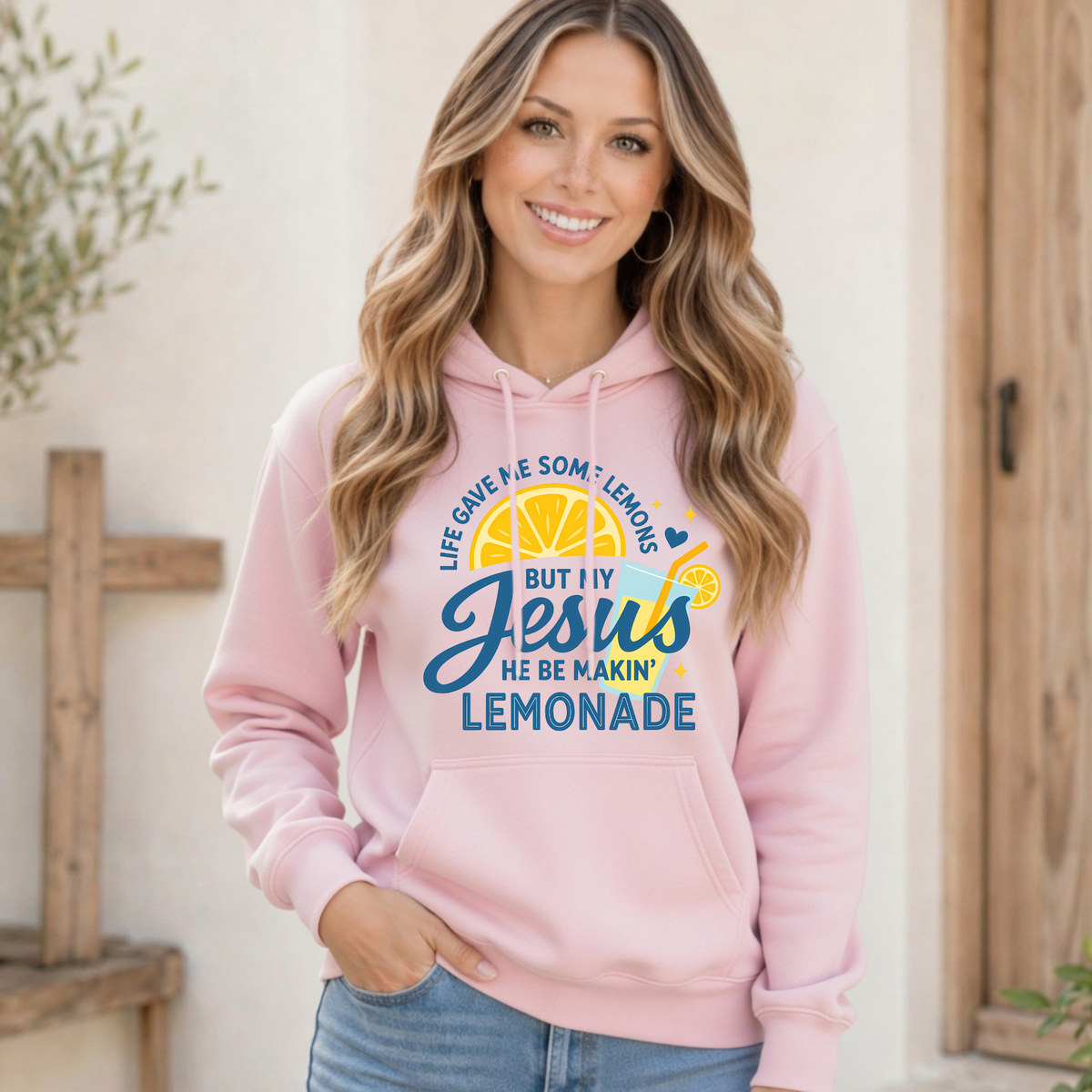 Smiling woman modeling a light pink hoodie featuring the Jesus Be Makin’ Lemonade lemon and drink design on a soft neutral backdrop.