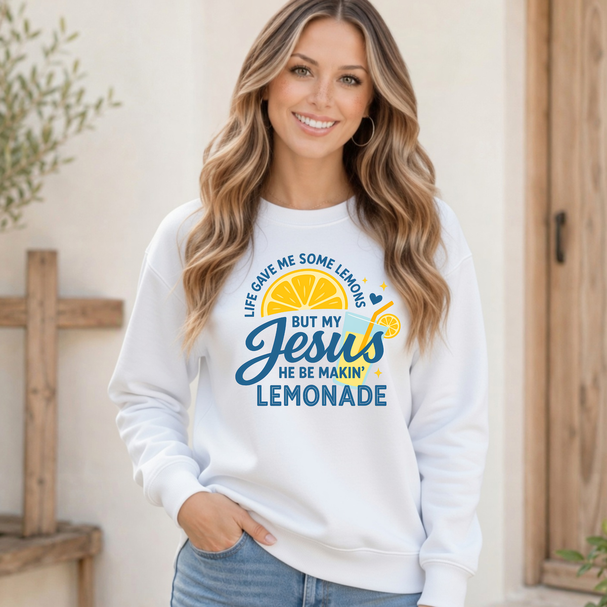 Woman wearing a white crewneck sweatshirt with the Jesus Be Makin’ Lemonade faith graphic featuring lemons, blue lettering and a lemonade cup.