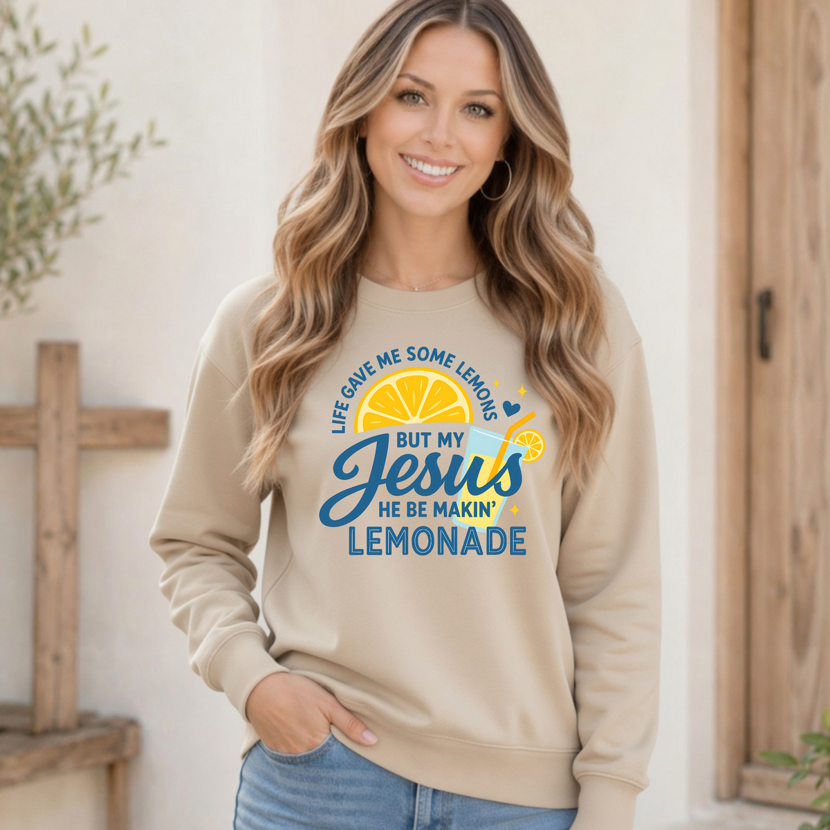 Woman wearing a sand crewneck sweatshirt featuring the Jesus Be Makin’ Lemonade graphic with bright lemon artwork and blue script lettering.