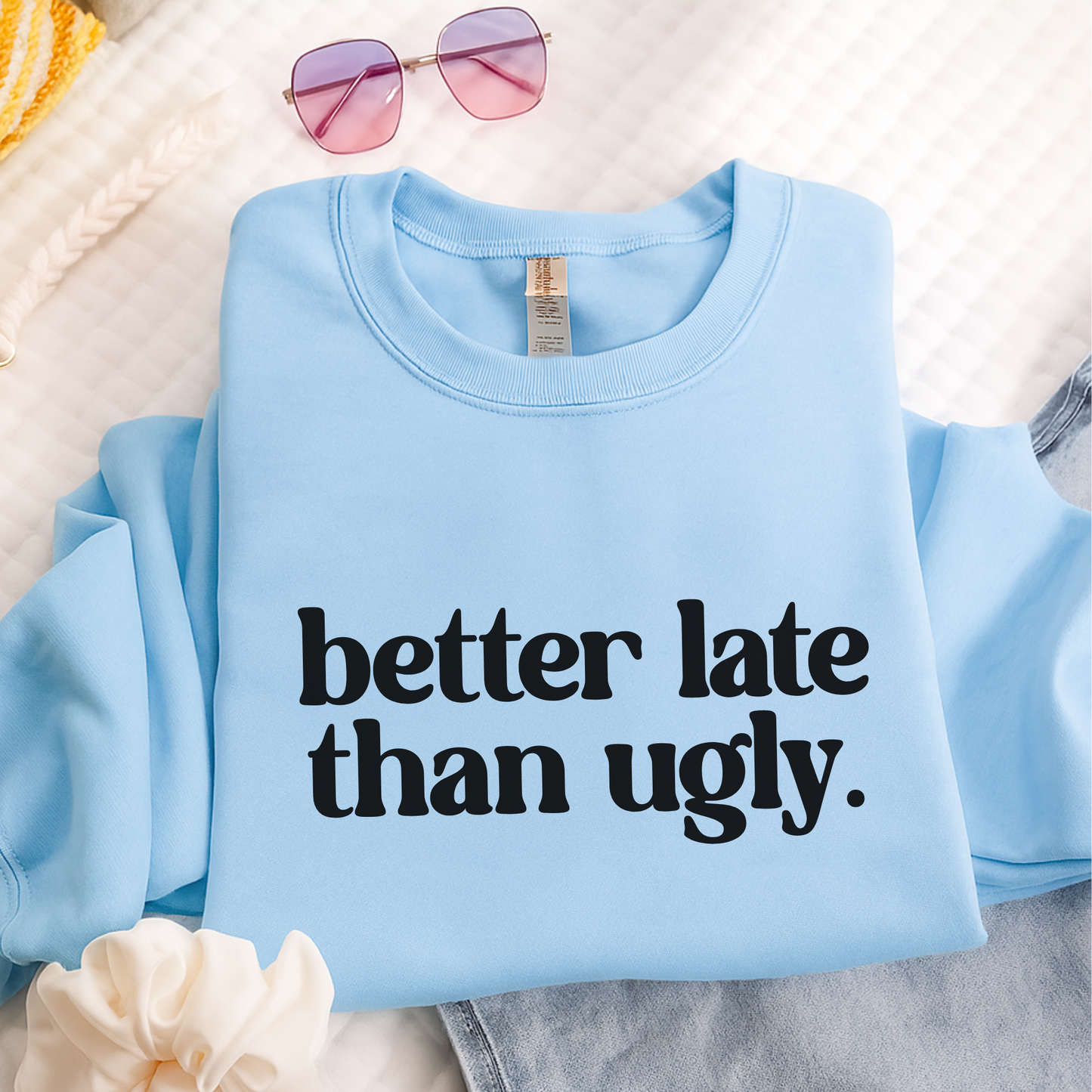 Light blue unisex crewneck sweatshirt with "Better Late Than Ugly" bold black graphic text. Folded on bed with jeans, sunglasses, and scrunchie for a trendy teen girl aesthetic. Fun and cozy pullover ideal for school or gifting.