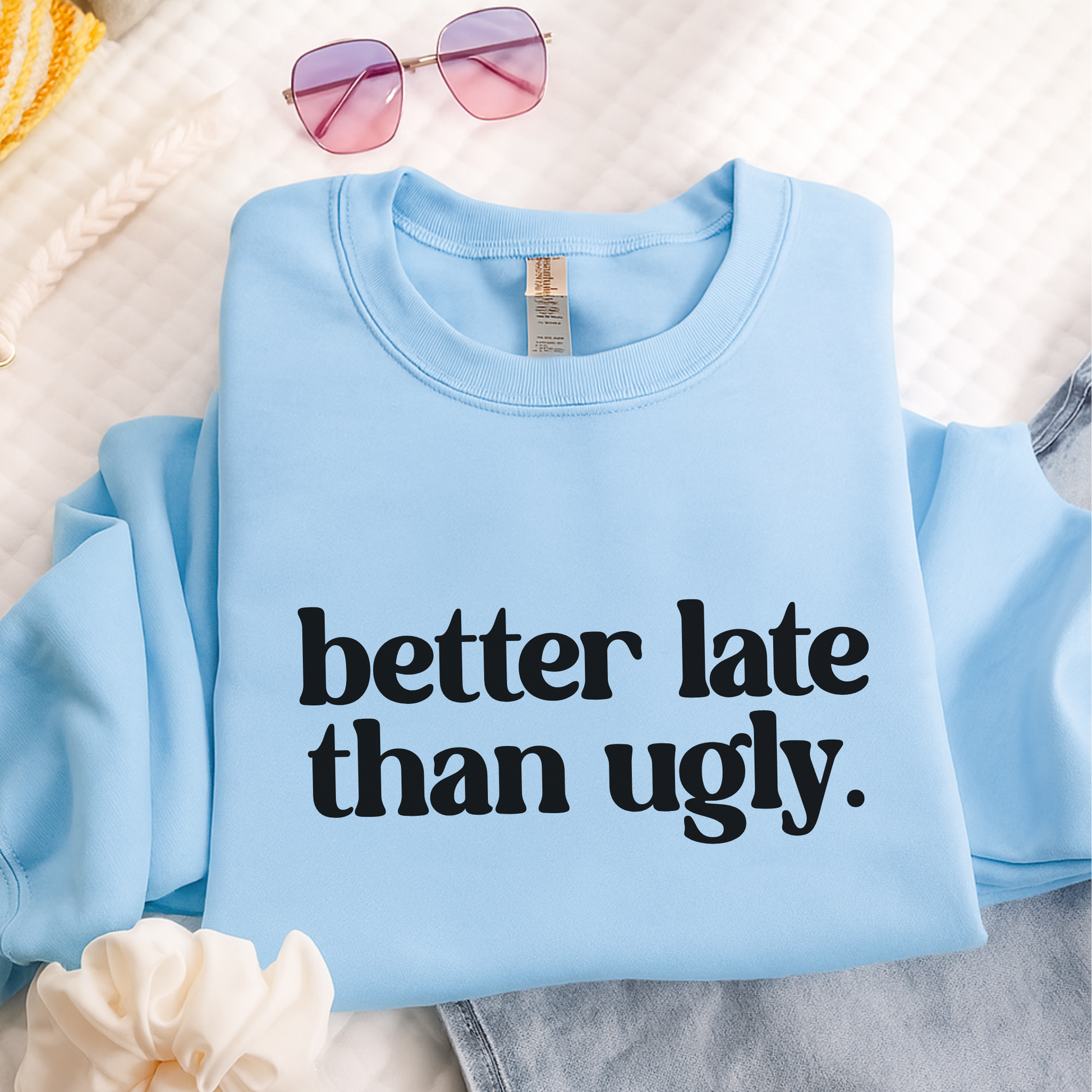 Light blue unisex crewneck sweatshirt with "Better Late Than Ugly" bold black graphic text. Folded on bed with jeans, sunglasses, and scrunchie for a trendy teen girl aesthetic. Fun and cozy pullover ideal for school or gifting.
