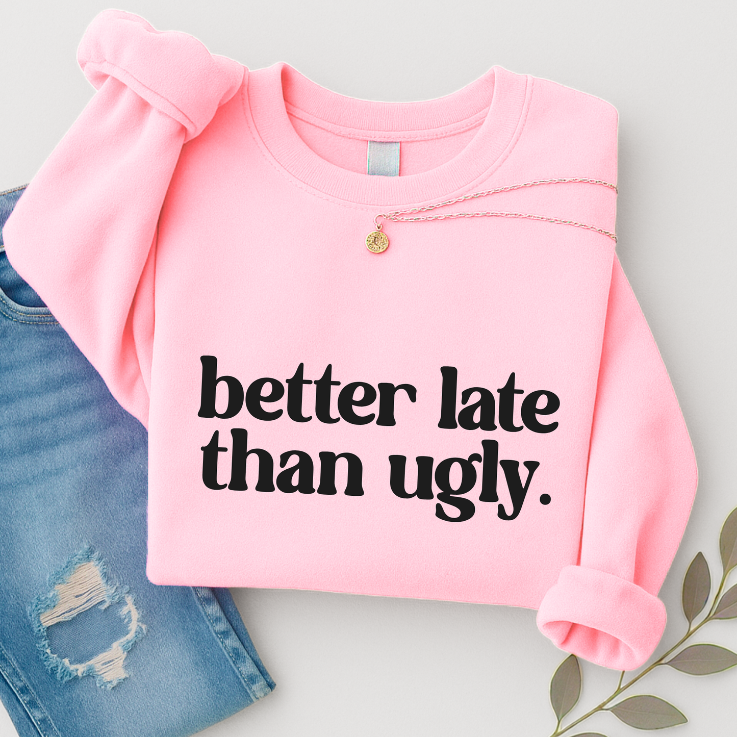 Light safety pink "Better Late Than Ugly" sweatshirt for women and teens, folded flat with sleeves rolled, styled with jeans and necklace. Trendy unisex pullover perfect for lounging, back-to-school, and casual outfits. Funny graphic crewneck for girls.