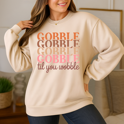 Light sand-colored sweatshirt with “Gobble Til You Wobble” in fall colors – woman smiling in cozy home setting, perfect for Thanksgiving.