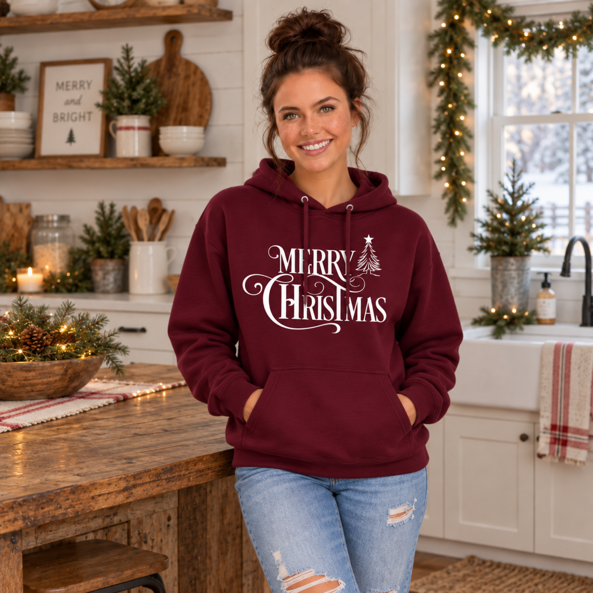 Maroon Merry Christmas hoodie on a model in a decorated kitchen, featuring a white script design and small Christmas tree.